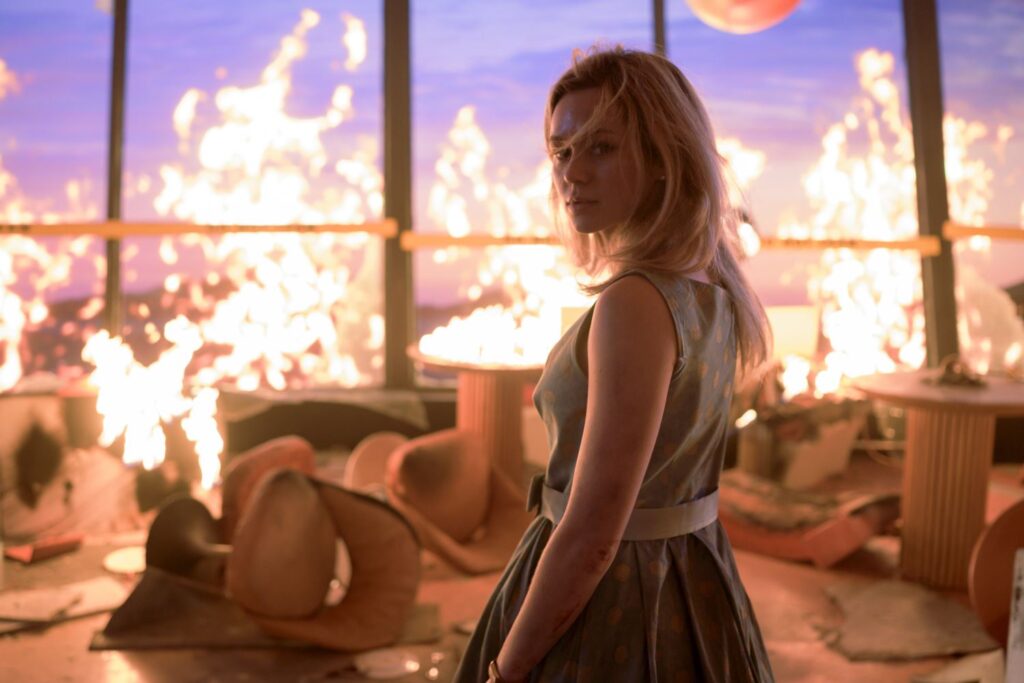 Brec Bassinger as “Iris” standing with flames behind her.