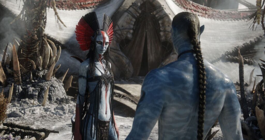 The fierce Mangkwan clan leader Varang, wearing a striking red and black headdress, stands facing away from the viewer towards a blue-skinned Na'vi character in a desolate, ash-filled environment.
