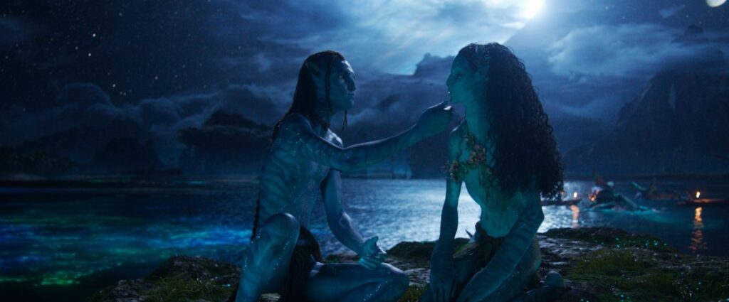 Lo'ak and Tsireya sitting by the water at night, gazing at each other with mountains in the background.