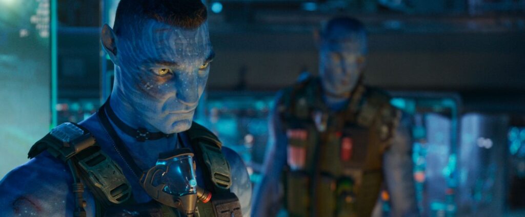 The recombinant character Miles Quaritch in the foreground and Lyle Wainfleet in the background, both tall blue humanoids wearing human military tactical vests inside a futuristic human base.