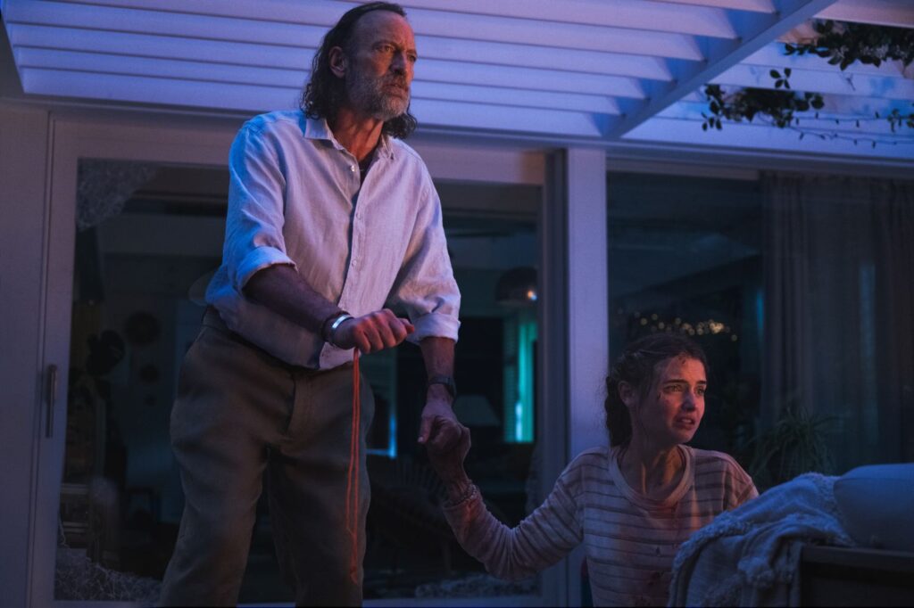 A man in a white shirt with his hands bound and a young woman covered in blood stare in horror from a house at night.