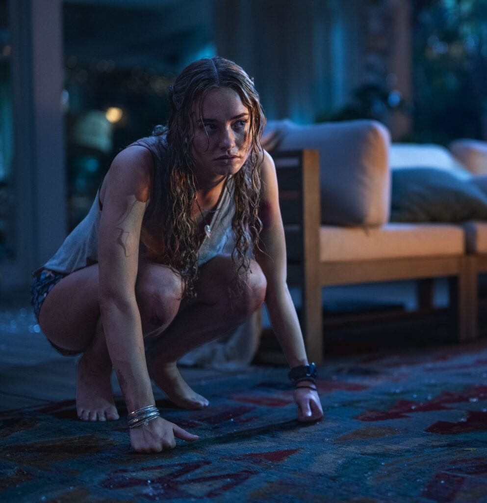 A young, wet woman with blood on her arms crouches in fear inside a house at night.