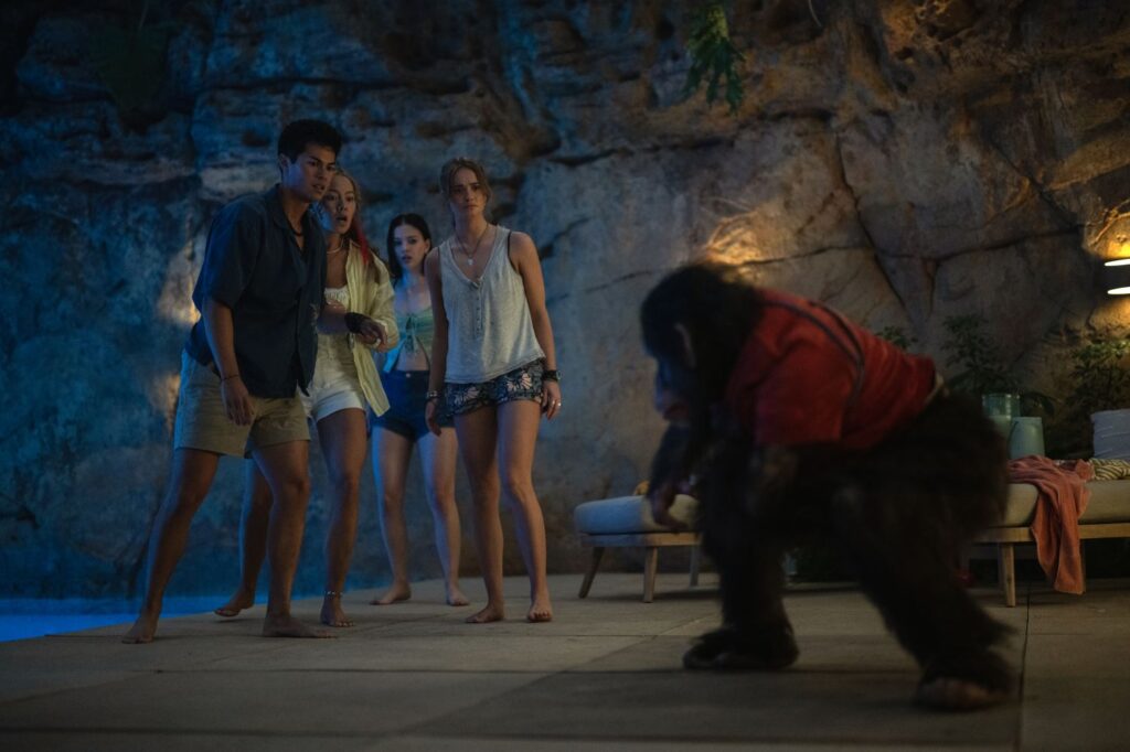 this film still from Primate is a group of four young adults reacting in horror to a rabid chimpanzee in a red shirt by a swimming pool in a tropical setting.