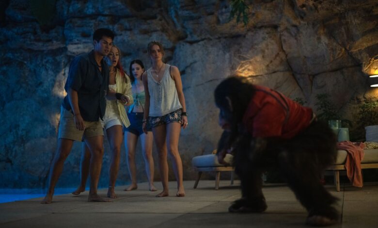 this film still from Primate is a group of four young adults reacting in horror to a rabid chimpanzee in a red shirt by a swimming pool in a tropical setting.