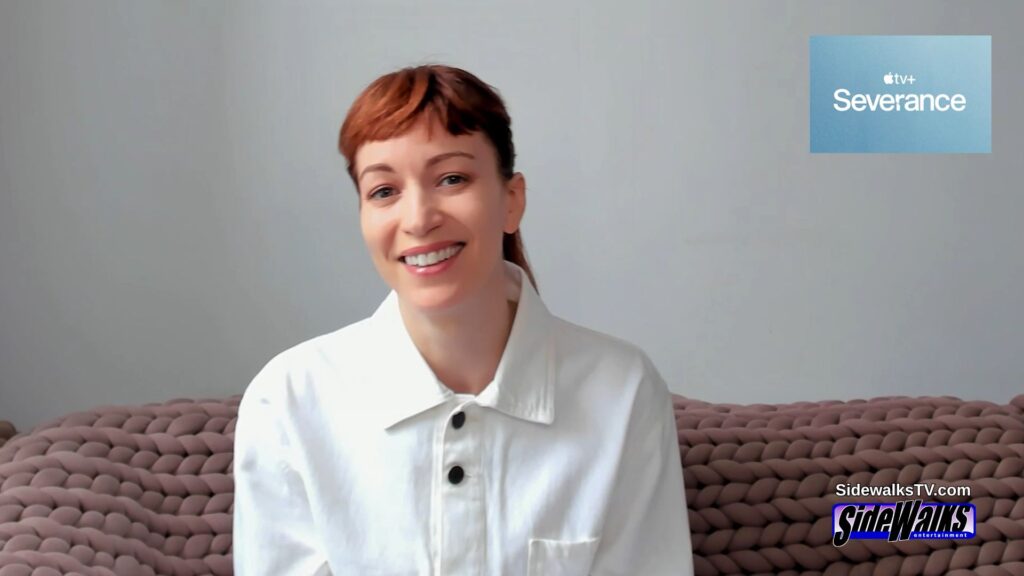 Gemini said Actress Britt Lower smiling during an interview for the Apple TV+ series "Severance". She wears a white shirt and sits before a chunky-knit sofa, with "Severance" and "Sidewalks Entertainment" logos visible.