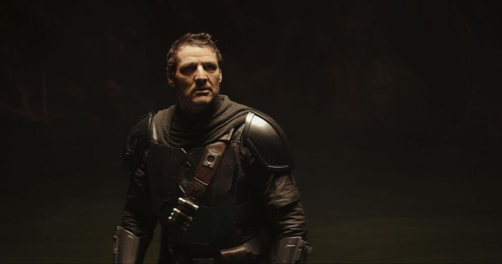 The Mandalorian (Pedro Pascal) is unmasked and standing and looking up in a scene.