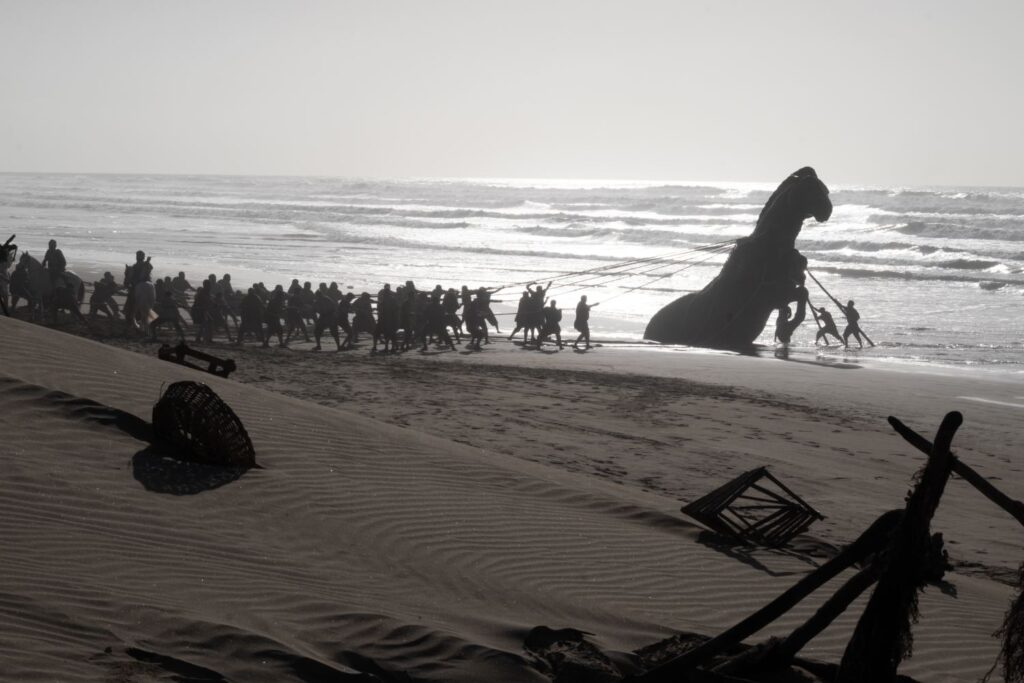 Silhouetted soldiers pulling a massive Trojan Horse structure onto a sandy beach from the ocean waves.